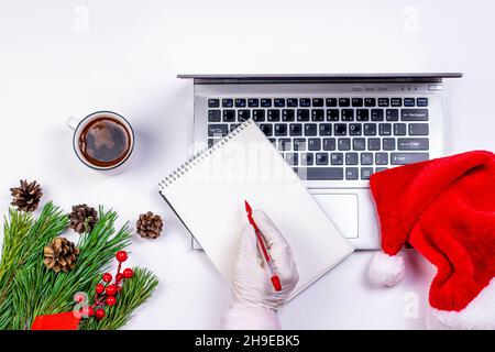 Weihnachtsmann-Hand mit Stift, Notizbuch und Laptop mit weihnachtlichen Dekorationen auf weißem Hintergrund. Alter Laptop mit Kaffeetasse, Santa`s Hut und Tannenzweig Stockfoto